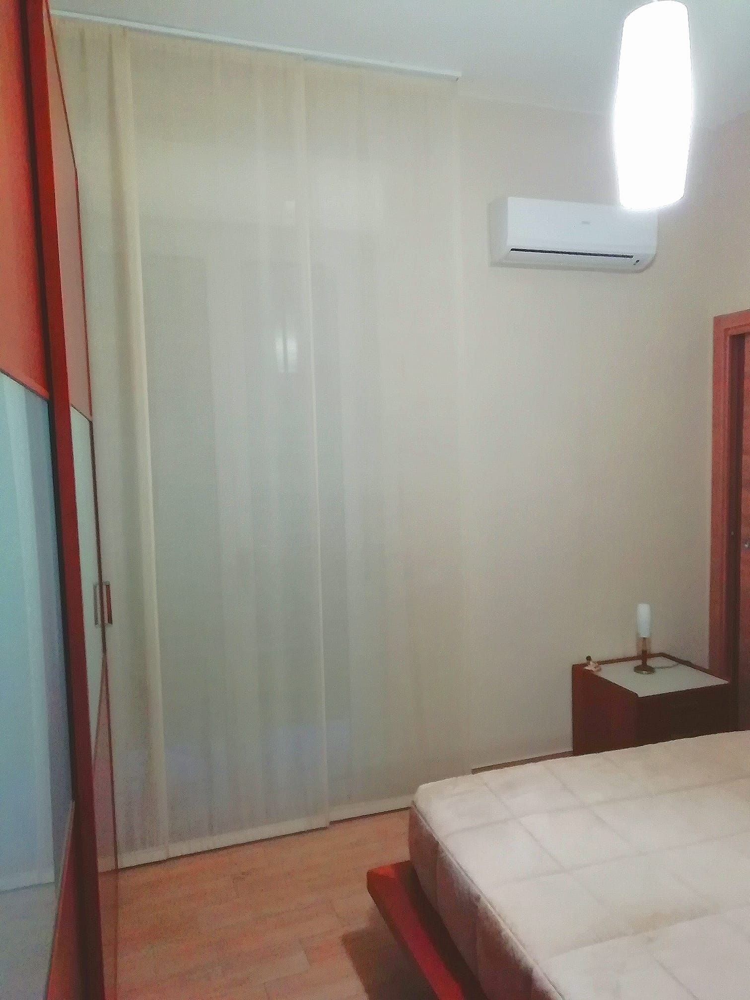 Piccola camera da letto con letto, comodino, condizionatore a parete e tende trasparenti a una finestra.
