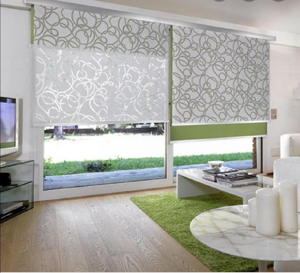 Soggiorno moderno con tende a rullo decorate, mobili bianchi e un tappeto verde vicino alle grandi finestre.