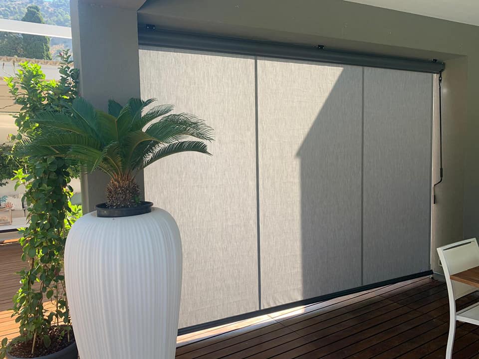 Patio esterno con grande fioriera bianca, pianta in vaso e tende beige chiuse accanto a una sedia bianca.