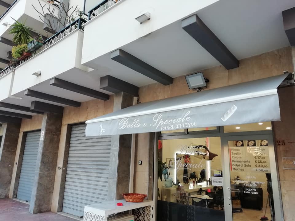 Negozio con vetrina su strada, tenda da sole, serrande metalliche e ingresso aperto in una fila di edifici.