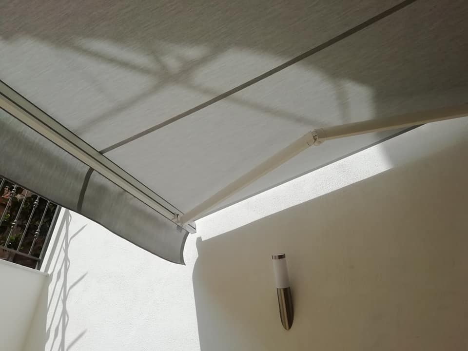 Una tenda da sole bianca proietta ombre su una parete beige illuminata dal sole, su cui è montata una piccola lampada a parete.