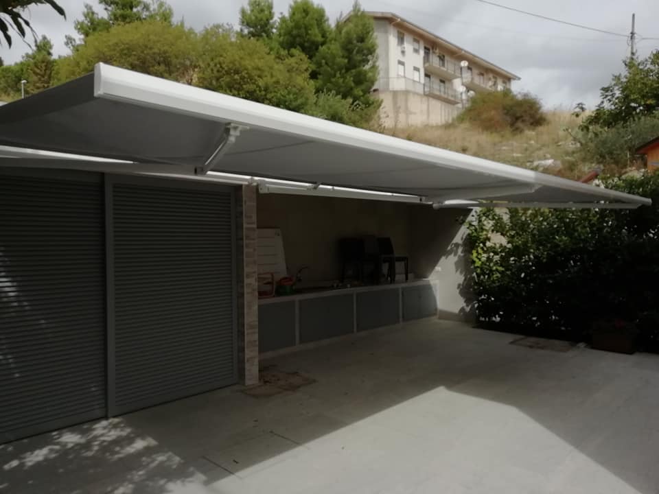 Posto auto coperto con tetto spiovente, porta metallica chiusa e vialetto in cemento accanto a una casa in collina.