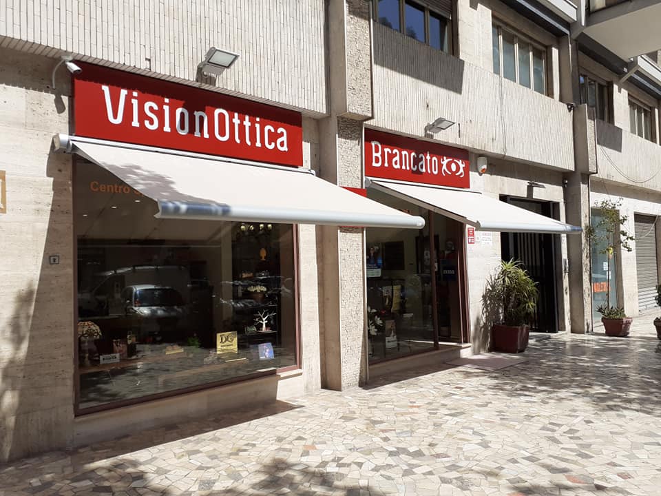 Vetrine di VisionOttica e Braccialini con insegne rosse e tende da sole bianche su un marciapiede soleggiato
