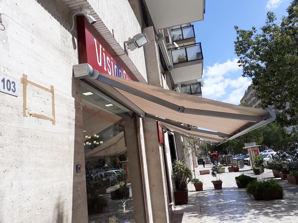 Negozio affacciato sulla strada con tenda da sole rossa, tettoia beige e piante in vaso lungo il marciapiede.