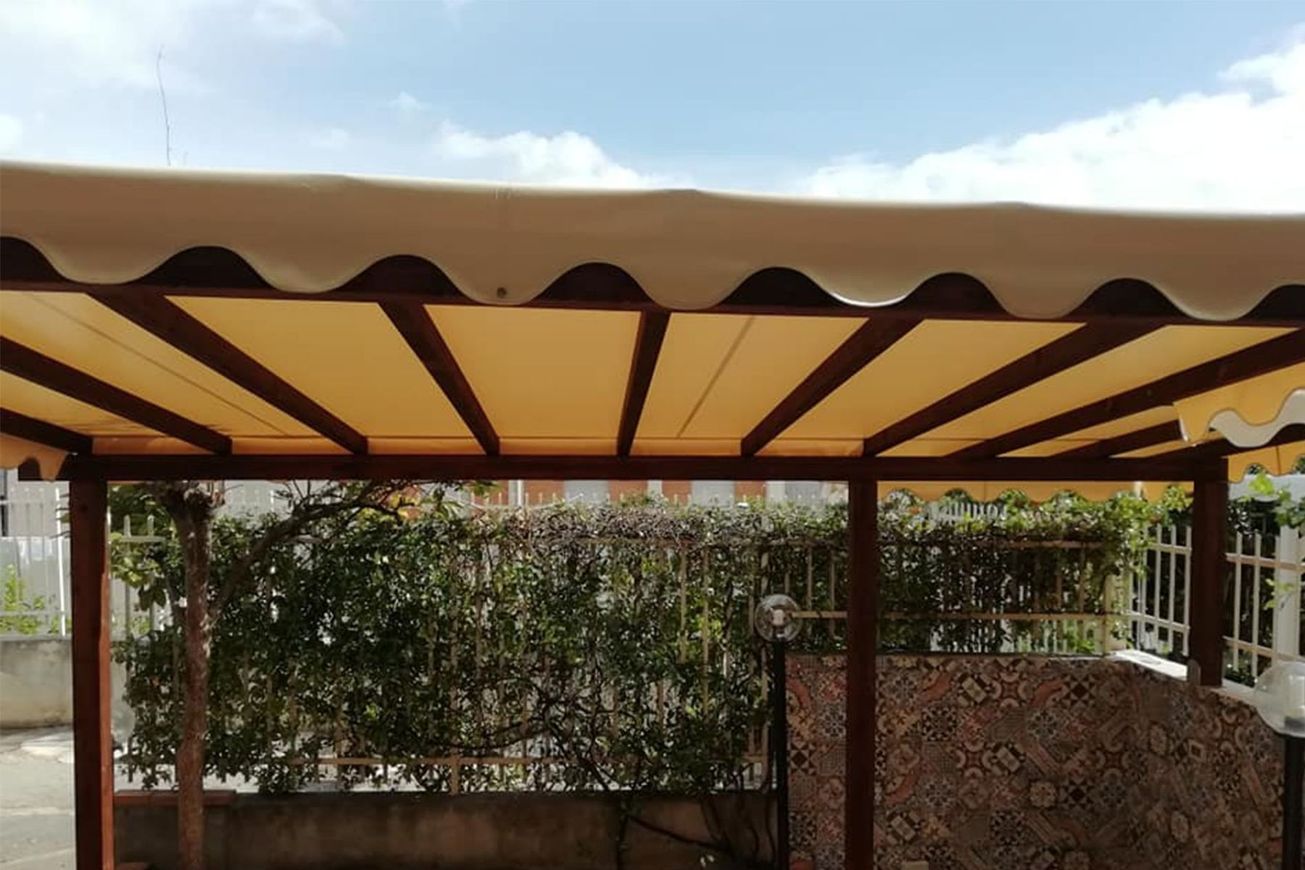 Tenda a righe beige e marroni sopra un patio con una siepe e un muretto decorativo in pietra sottostante.