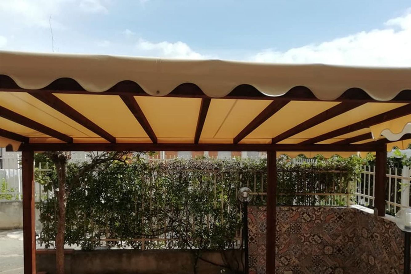 Tenda a righe beige e marroni sopra un patio con una siepe e un muretto decorativo in pietra sottostante.