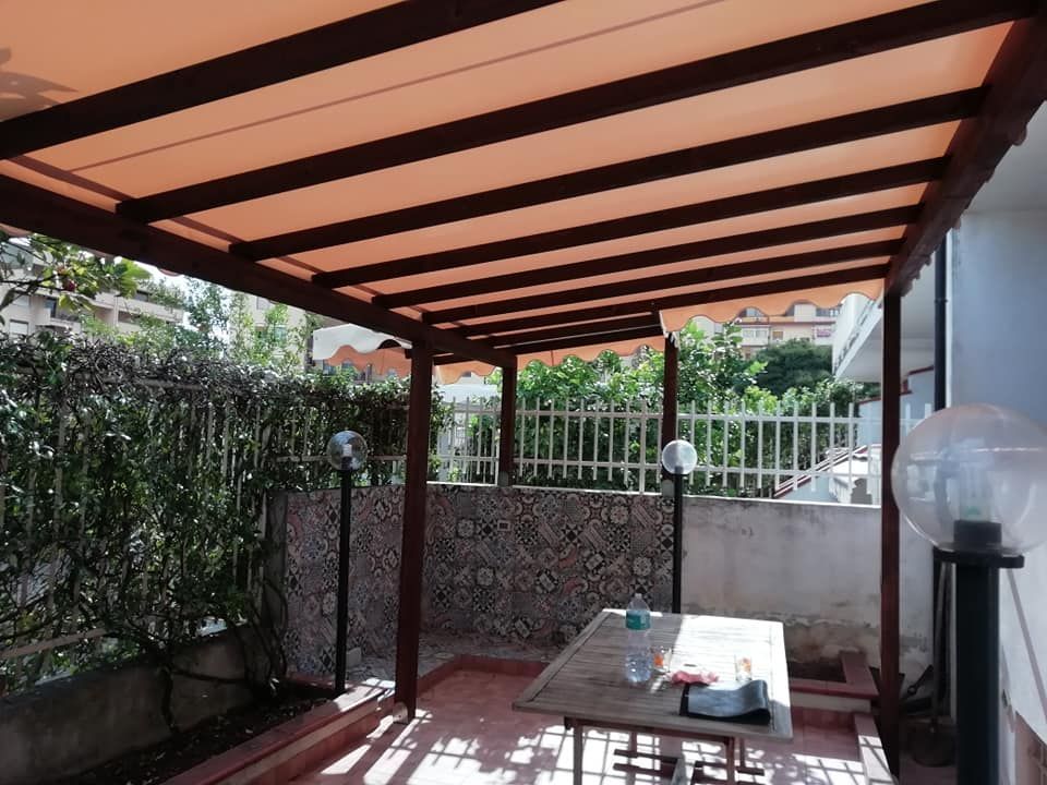 Patio coperto con travi in ​​legno, pavimento piastrellato e un tavolino accanto a una recinzione da giardino.