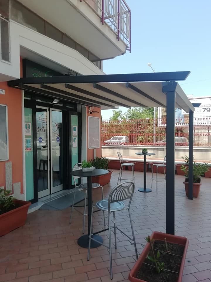 Dehor esterno di un caffè con tavoli, sedie e tende da sole nere, adiacente a una vetrina.