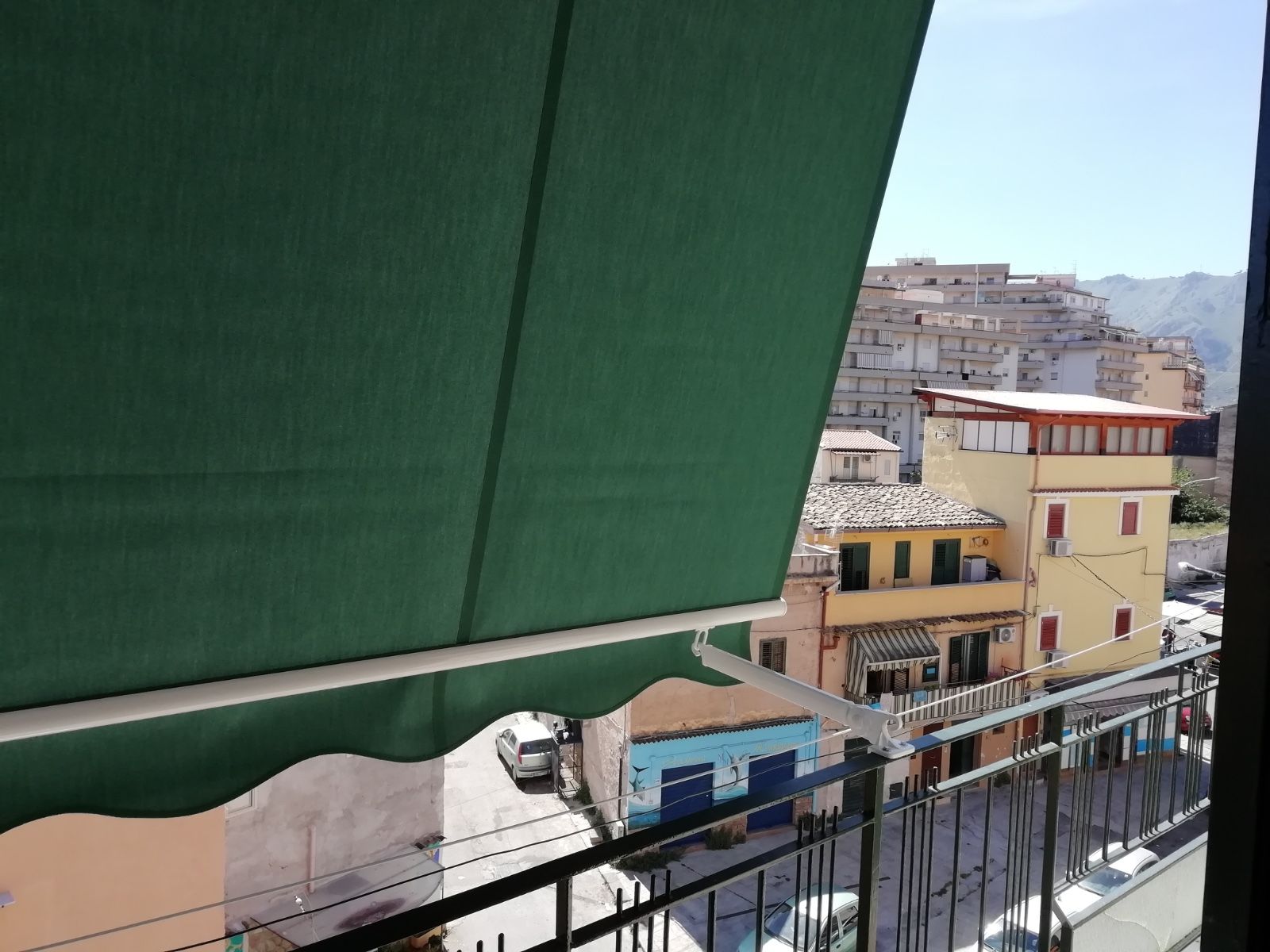 Tenda verde affacciata su una soleggiata strada cittadina con palazzi e auto parcheggiate.