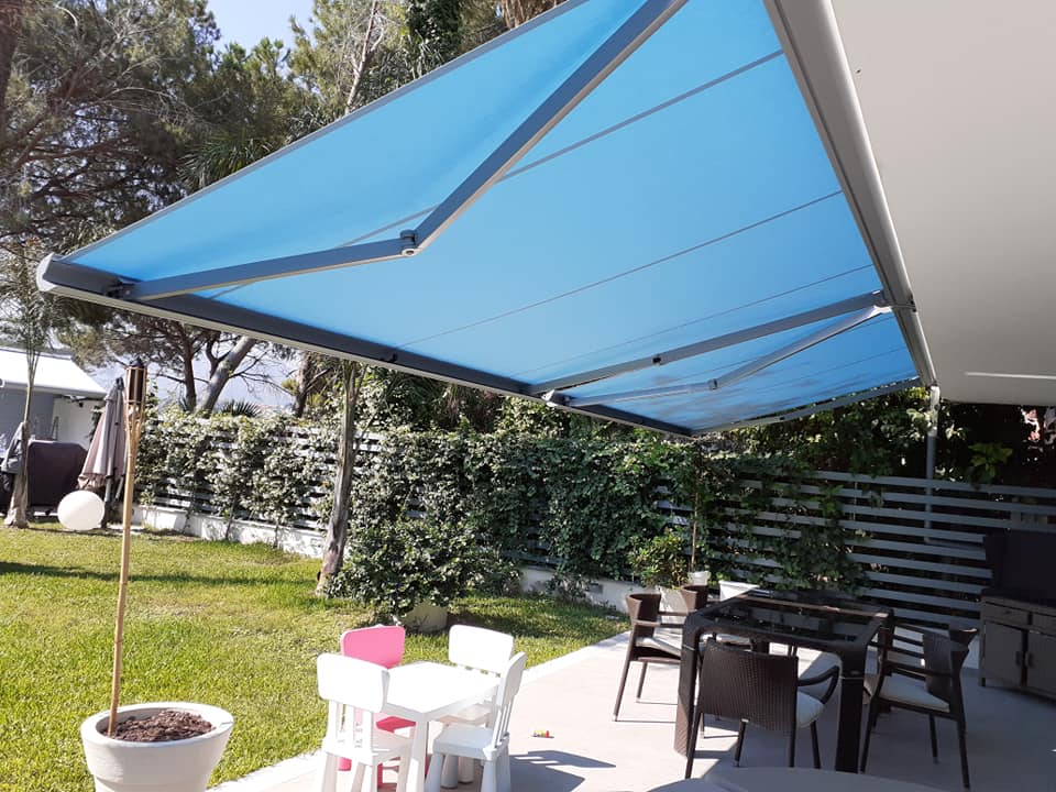 Una tenda da sole blu ombreggia un patio esterno con sedie, tavolo e un prato adiacente.