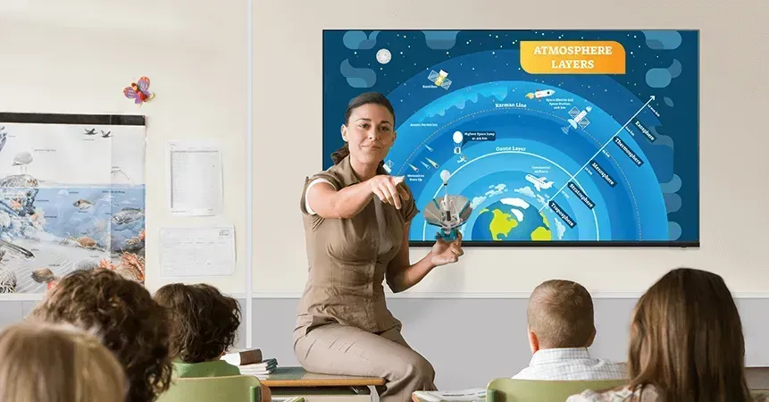 Teacher pointing at a globe model while explaining a diagram about atmospheric layers to children in a classroom.