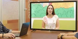 A person on a screen converses with two people at a table, one using a laptop.