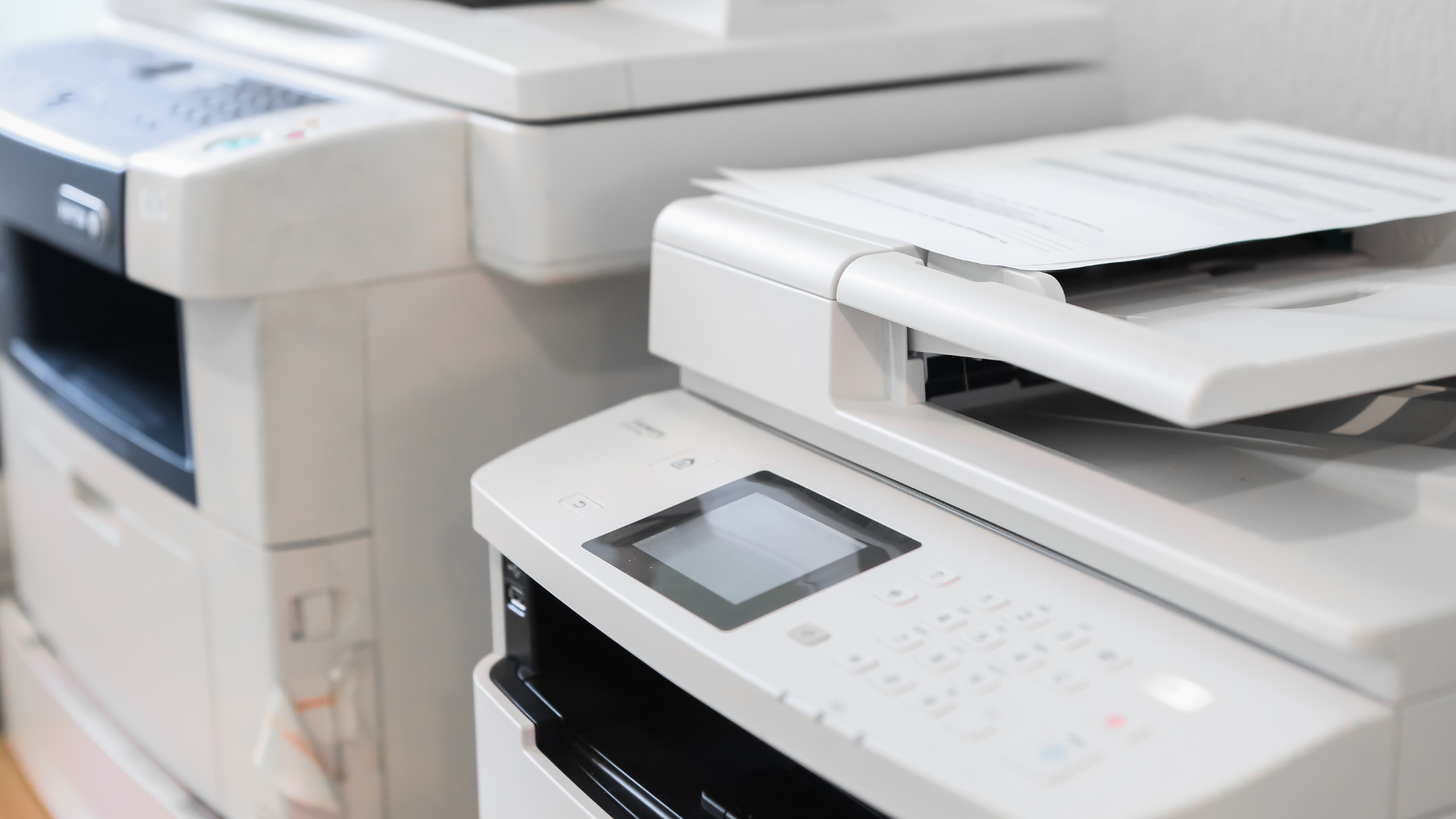 Two white office printers with open paper trays, sitting side-by-side.