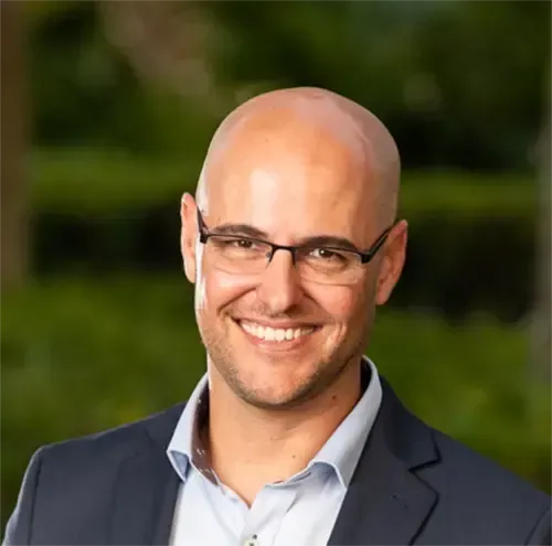 Bald man with glasses smiling, wearing a blue blazer and open-collared shirt, against a green background.