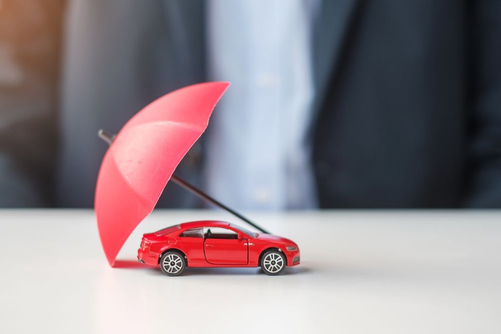Red toy car protected by a small red umbrella, symbolizing insurance. A person in a suit is in the background.