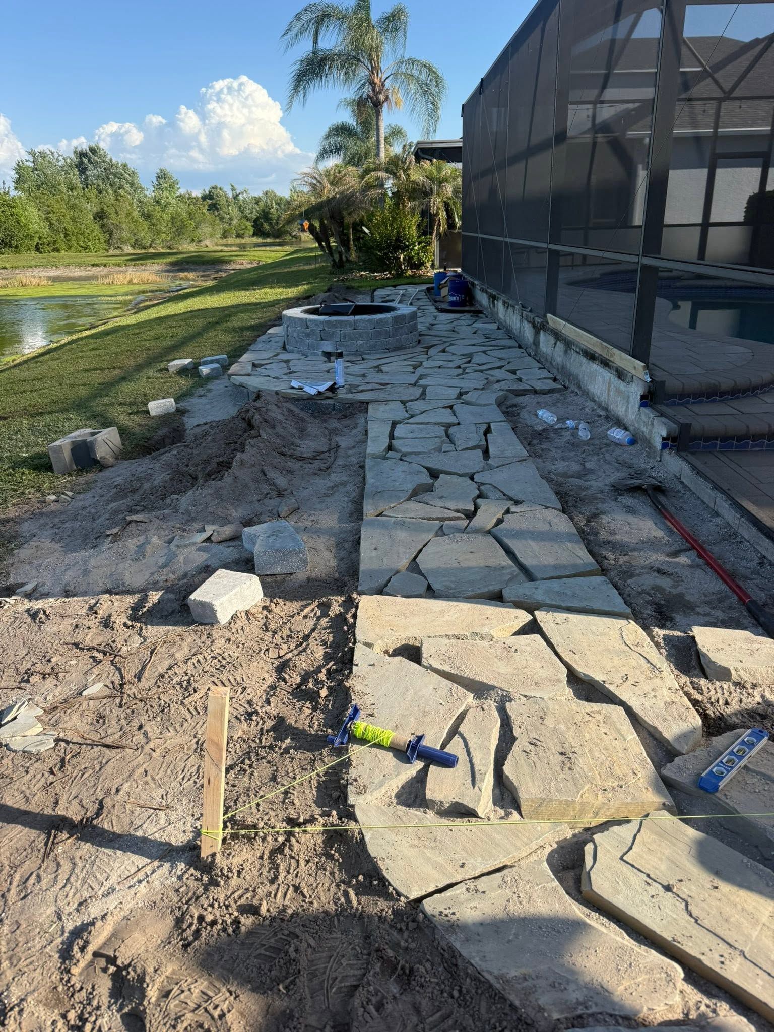 Stone walkway under construction beside a screened building, with tools and a pond in a sunny yard.