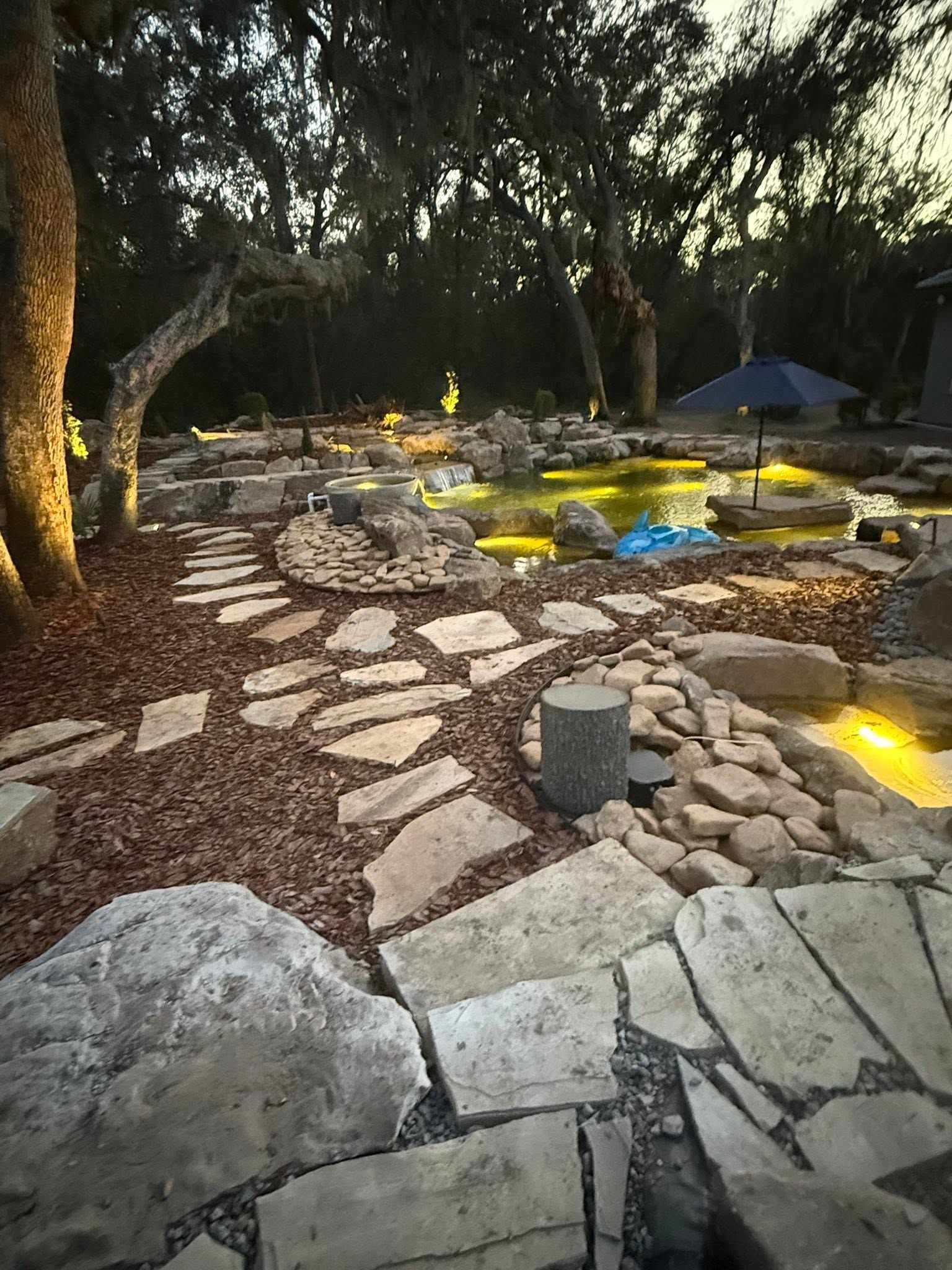 Stone path and illuminated garden seating under large trees at dusk