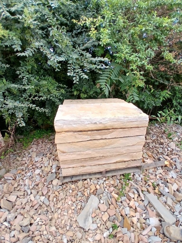 Stack of light tan paving stones on gravel, with dense green bushes in the background