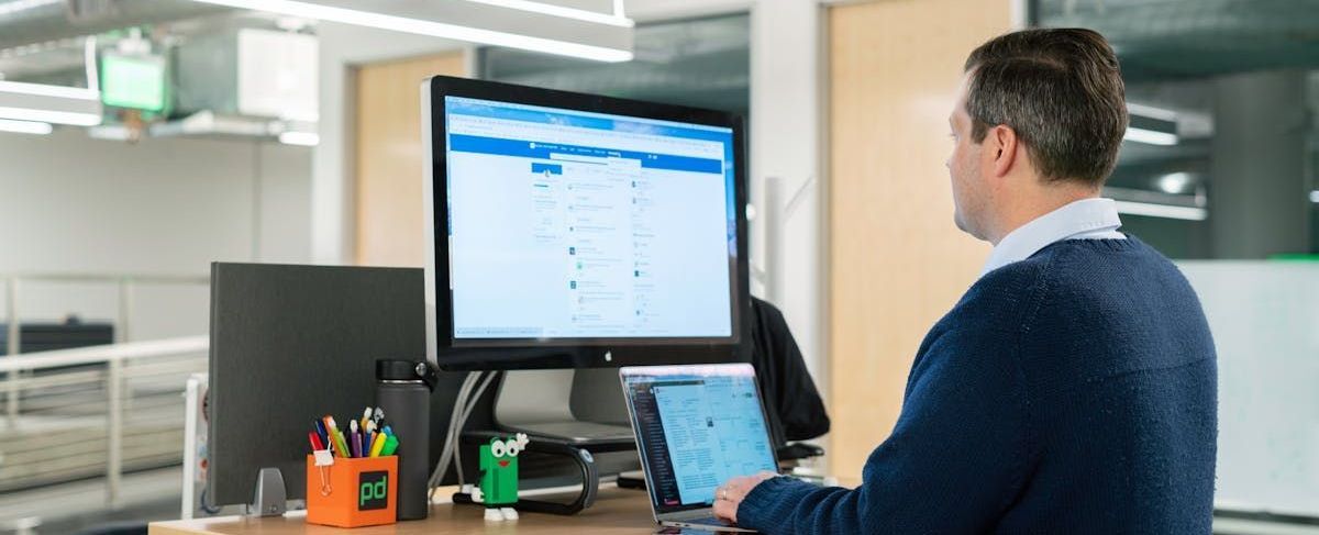 Um homem está sentado em uma mesa em frente a um computador e um laptop.