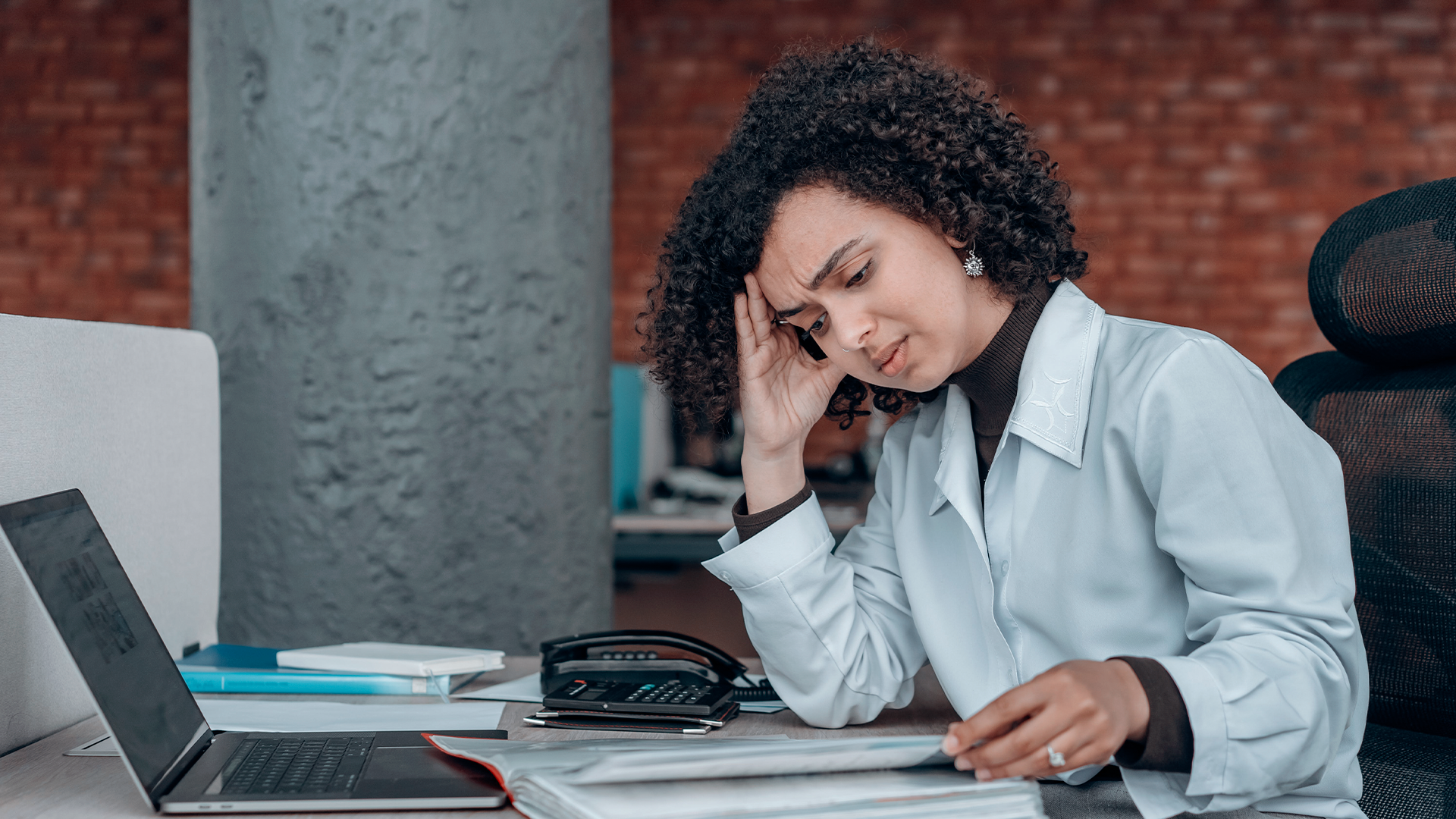 Uma mulher tensa por causa da Síndrome de burnout  sentada  com um laptop e papéis a sua frente.