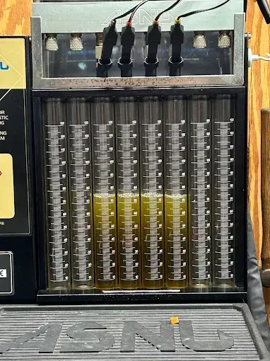 Fuel injector testing machine displaying fuel flow in clear tubes. Tubes contain yellow liquid.