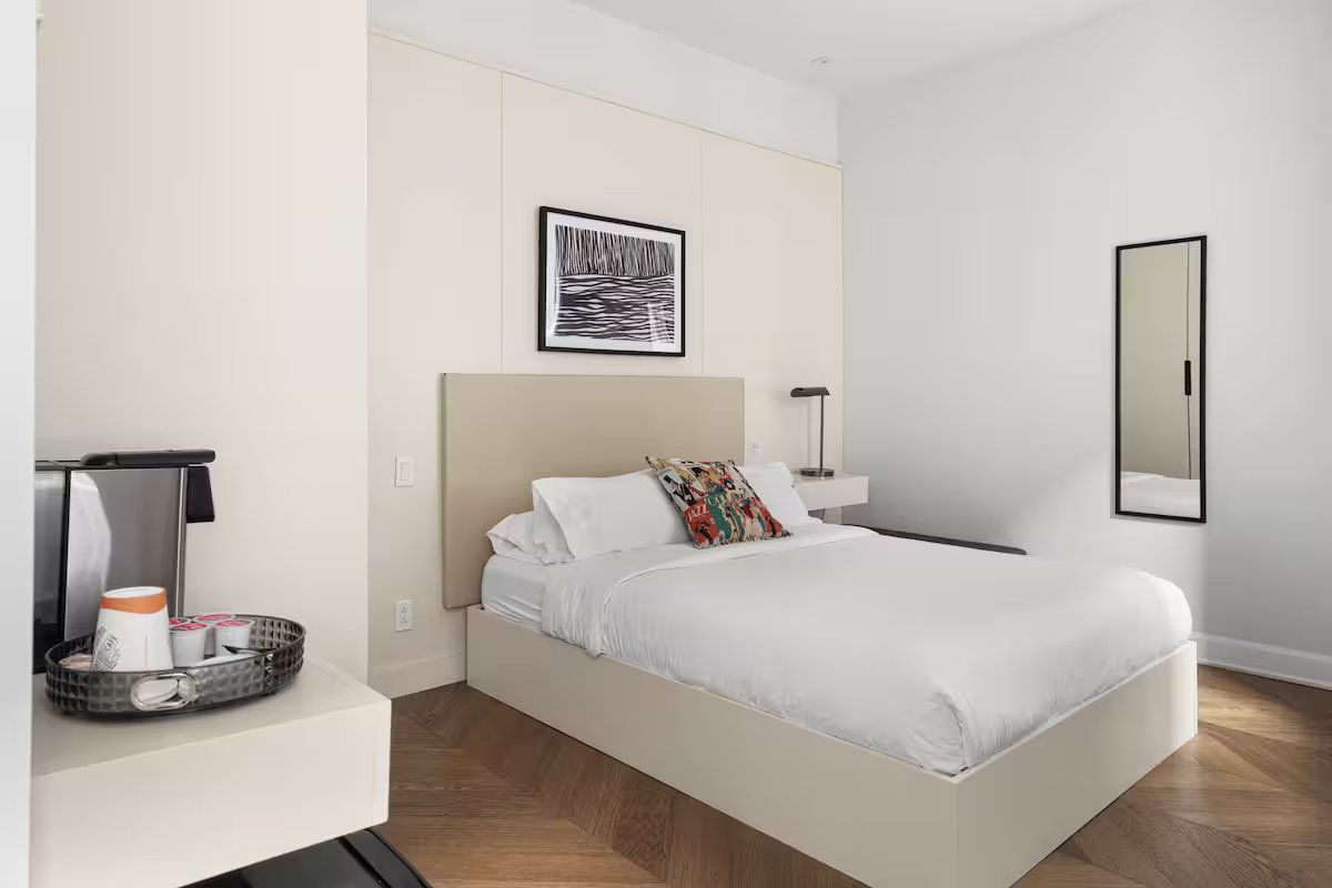 A hotel bedroom with a bed, small side table, mirror, and coffee station. The room is light and airy.