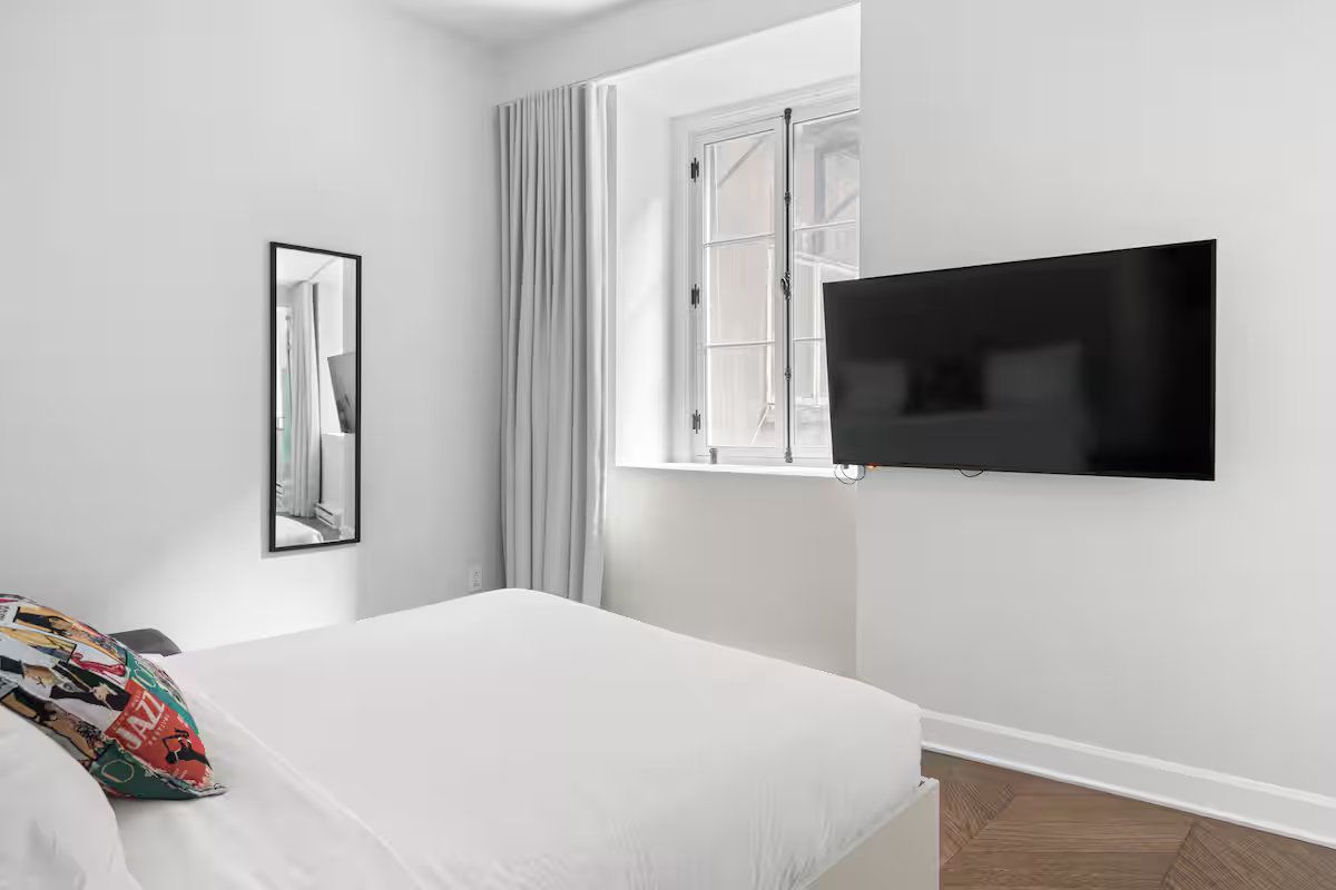 A modern, minimalist bedroom with a bed, a mounted television, a floor-length mirror, and white curtains by a window.