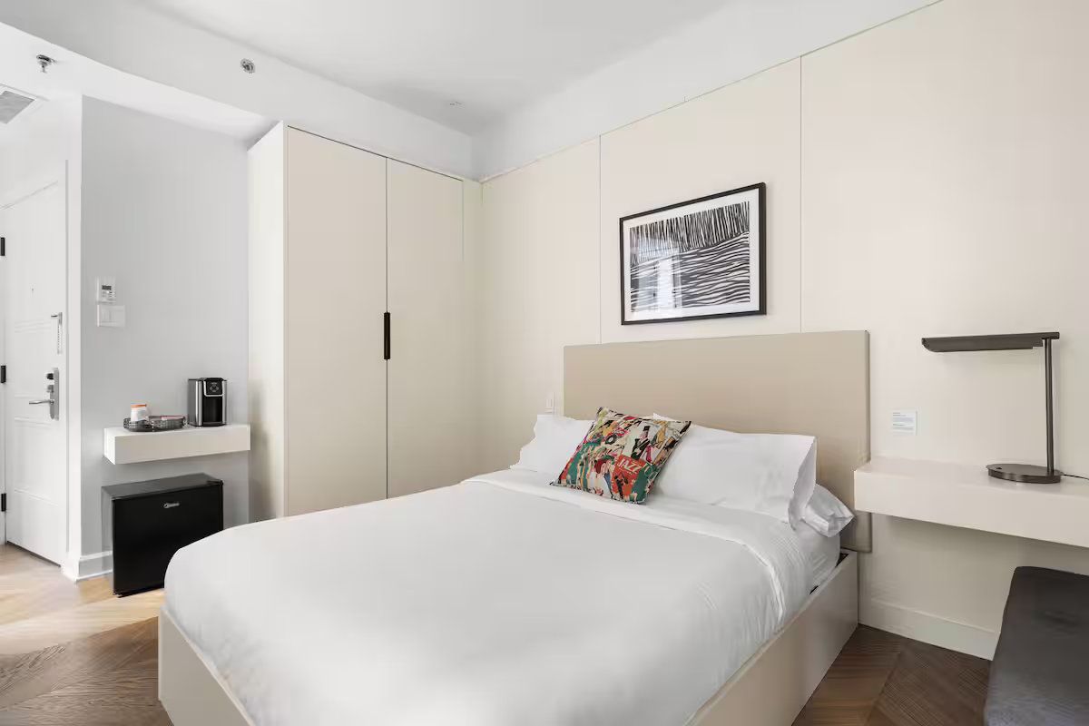 A modern, minimalist bedroom with a white bed, beige headboard, large wardrobe, and wall-mounted desks on light wood floors.
