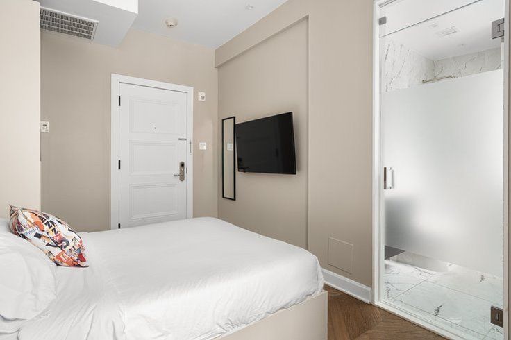 Bedroom with a bed, TV, closed door, and glass-enclosed shower. Walls are beige, floor is wood.