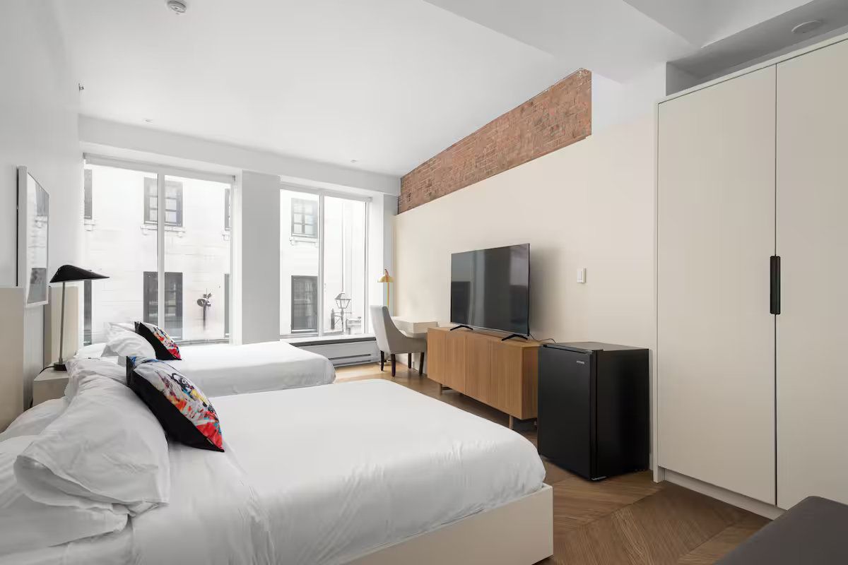 Bright hotel room with two beds, large windows, TV, and wardrobe.