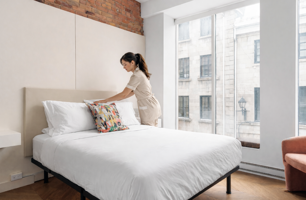 Woman in uniform making a bed, placing a white blanket. Bedroom with a nightstand and lamp.