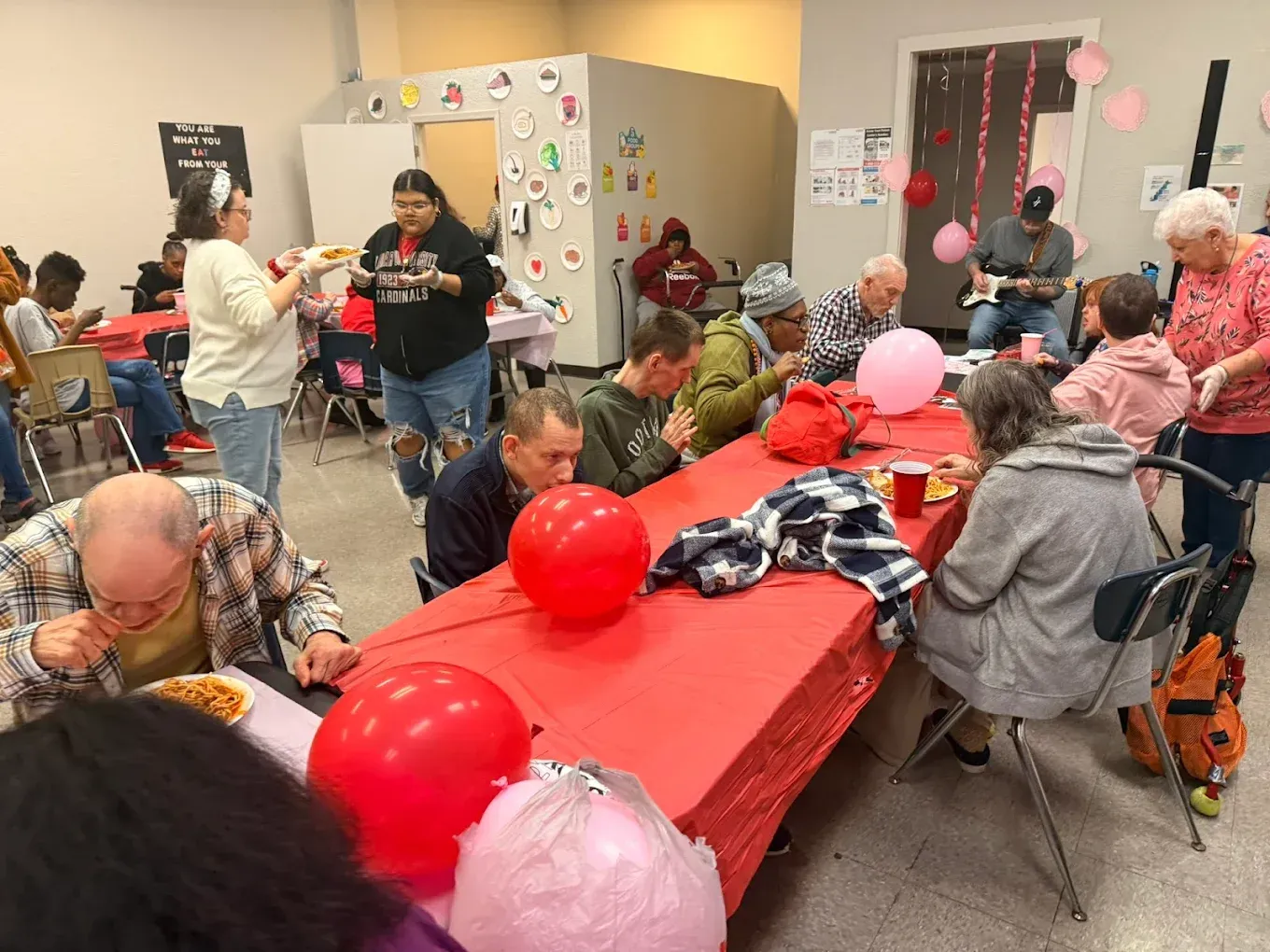 A group of people are sitting at long tables with red tables cloths and pink balloons.