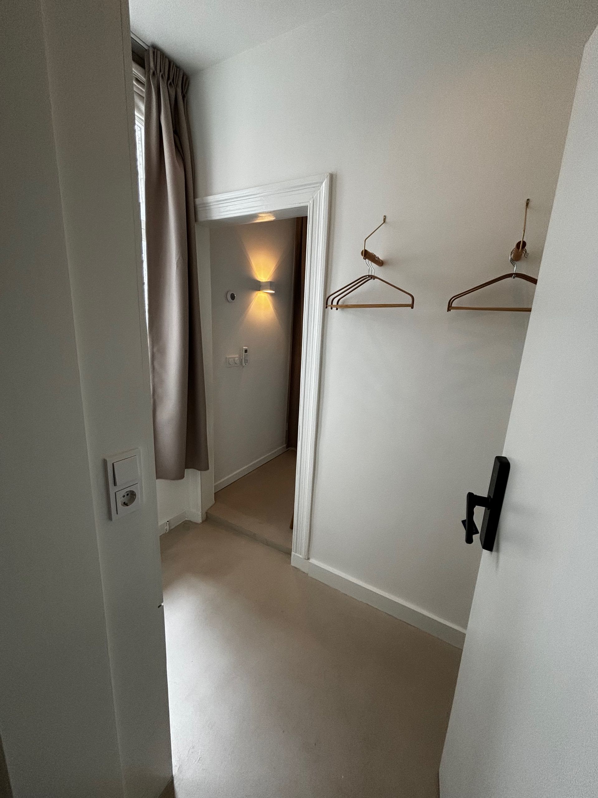 Gang met beige vloerbedekking, witte muren, open deur rechts en twee kledinghangers.