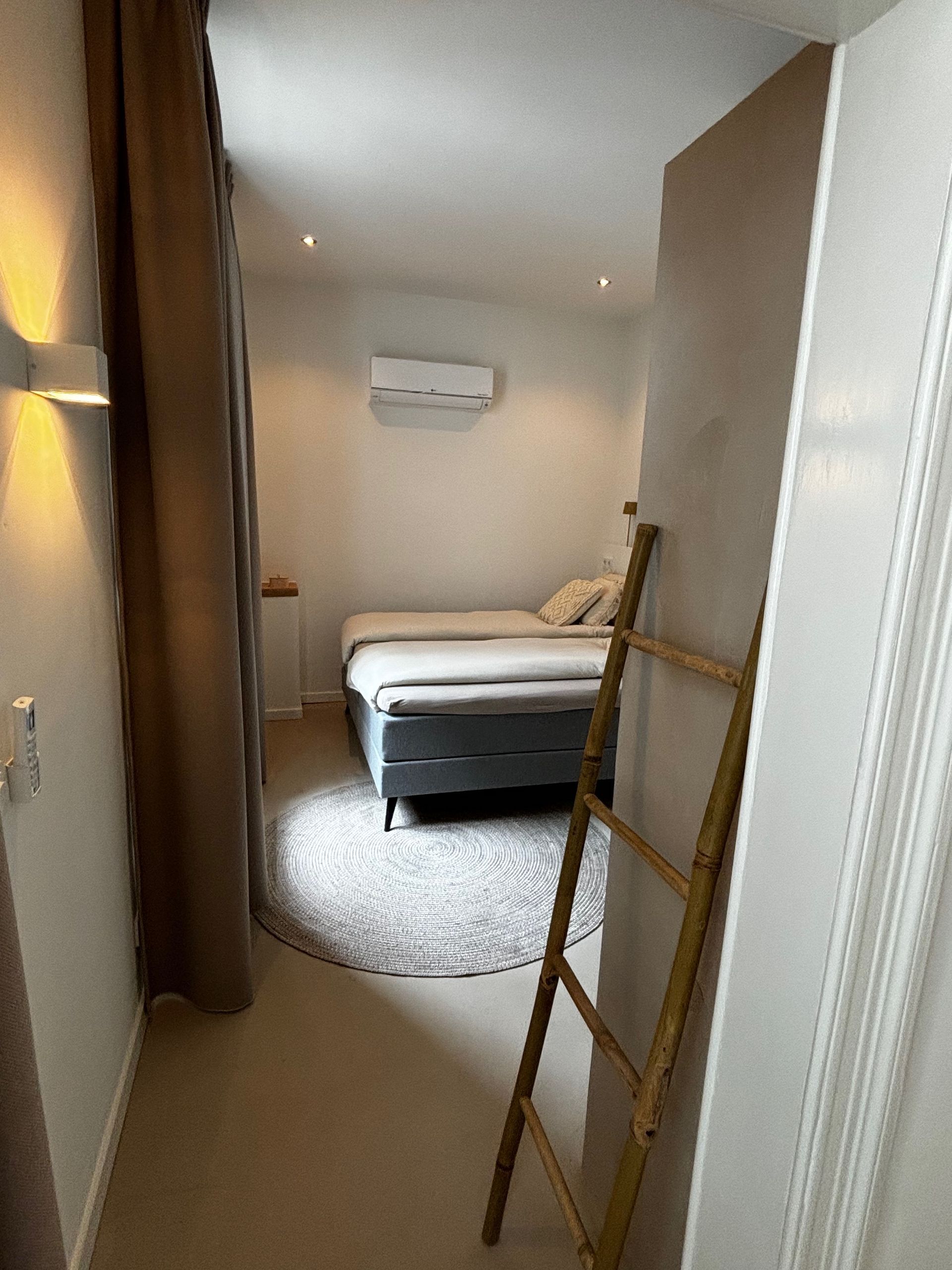Smalle gang die naar een slaapkamer leidt. Een bed is zichtbaar met een rond vloerkleed en een decoratieve ladder.