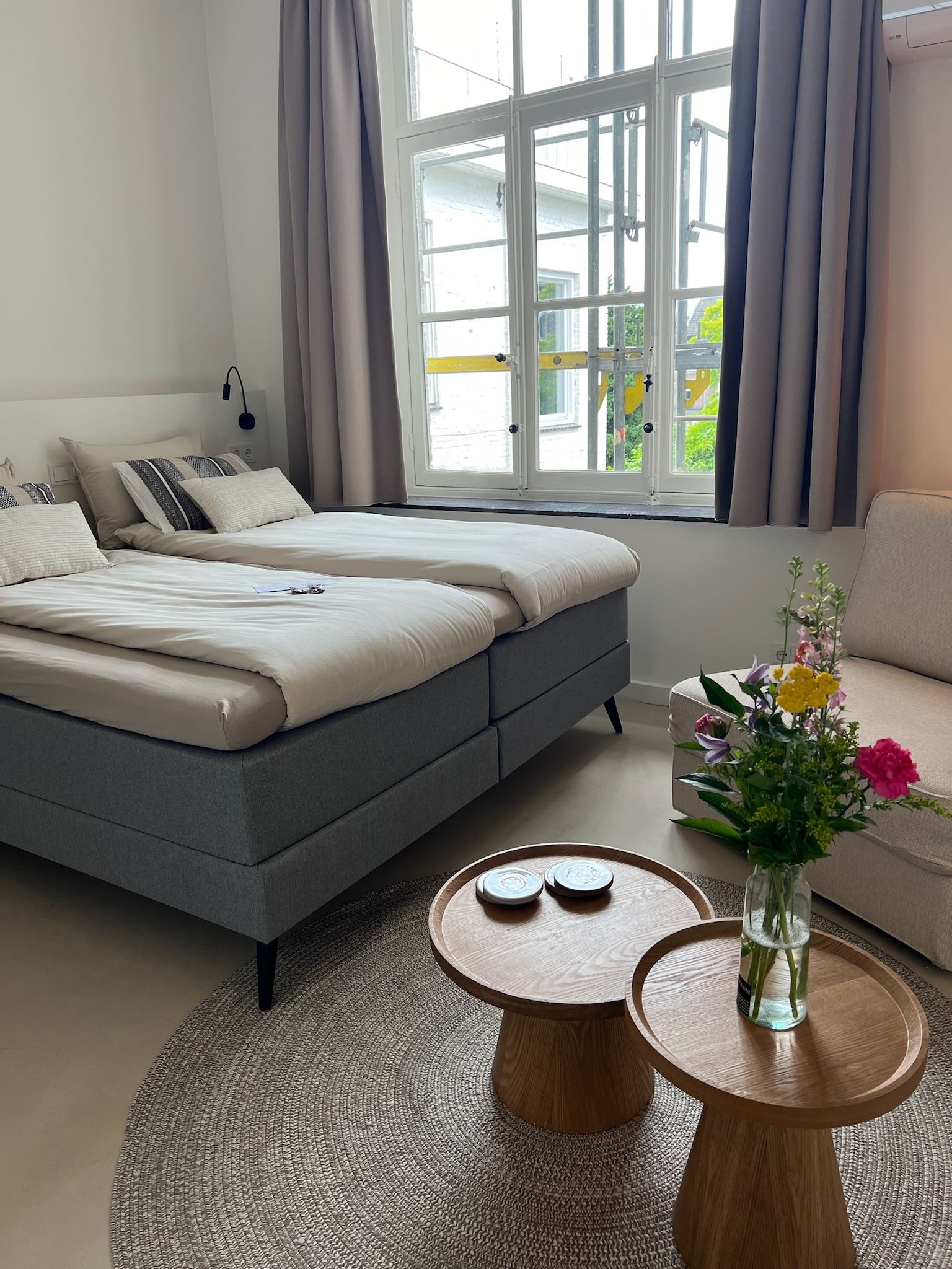Slaapkamer met twee bedden, kleine tafeltjes op een kleed, bloemen en een raam met gordijnen.