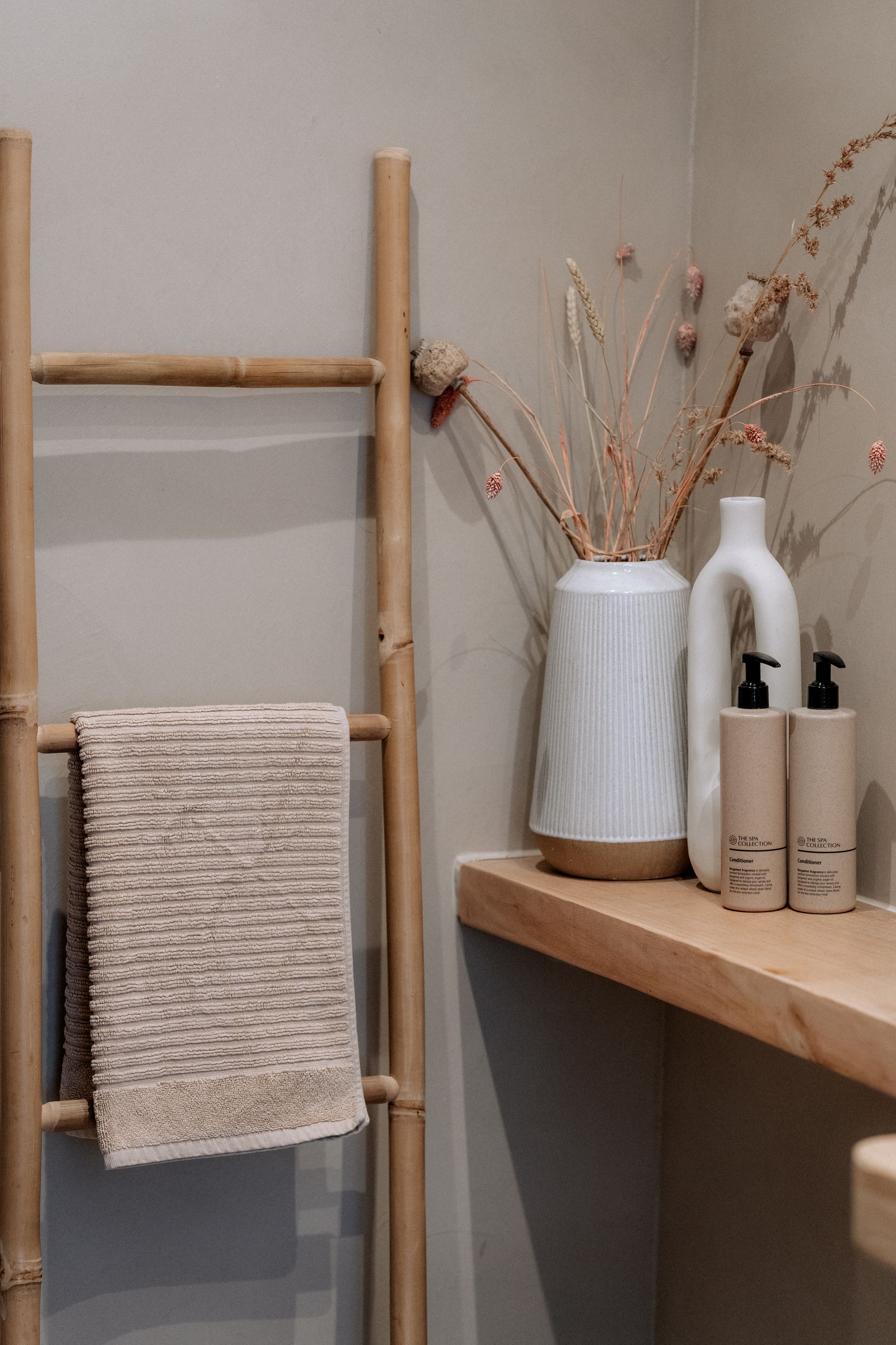 Een handdoekrek van bamboe in de badkamer. Een vaas met gedroogde bloemen en toiletartikelen.