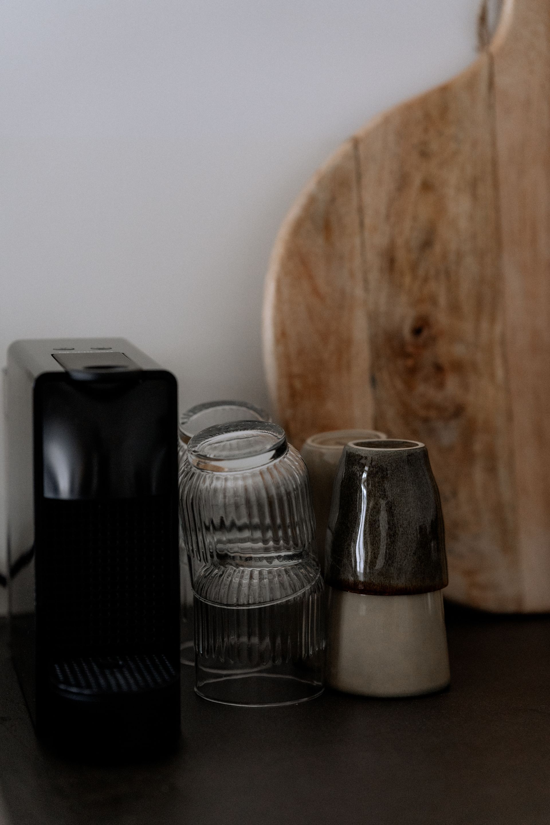 Een zwart koffiezetapparaat, opgestapelde glazen en een houten snijplank op een donkere achtergrond.