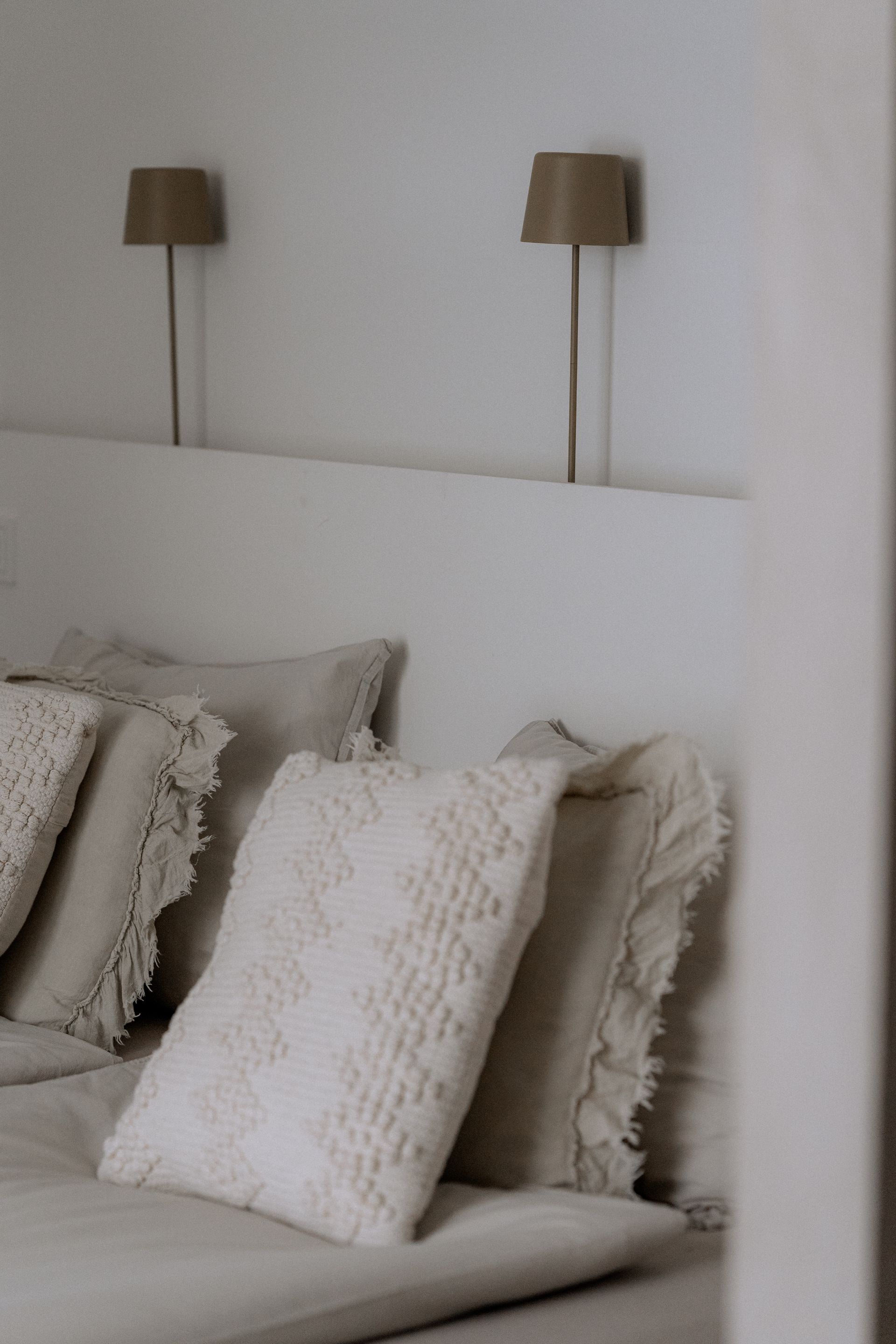 Een bed met crèmekleurige kussens en wandlampen in een slaapkamer met neutrale tinten.
