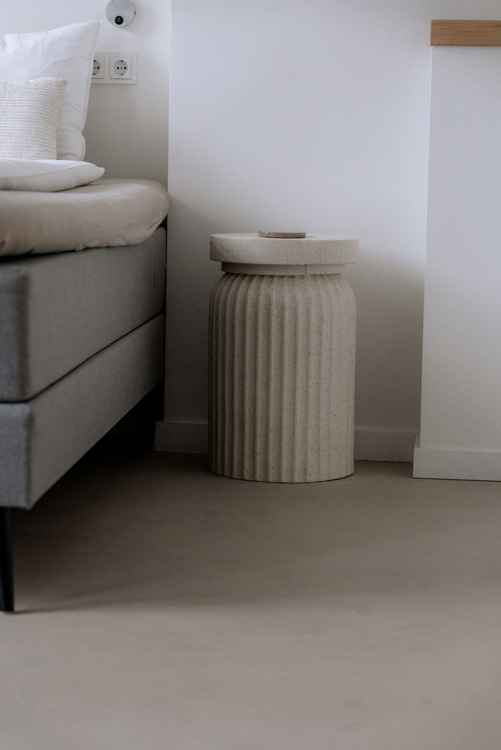 Beige bijzettafel met ribbels naast een bed met een grijze basis en witte kussens, in een witte kamer.