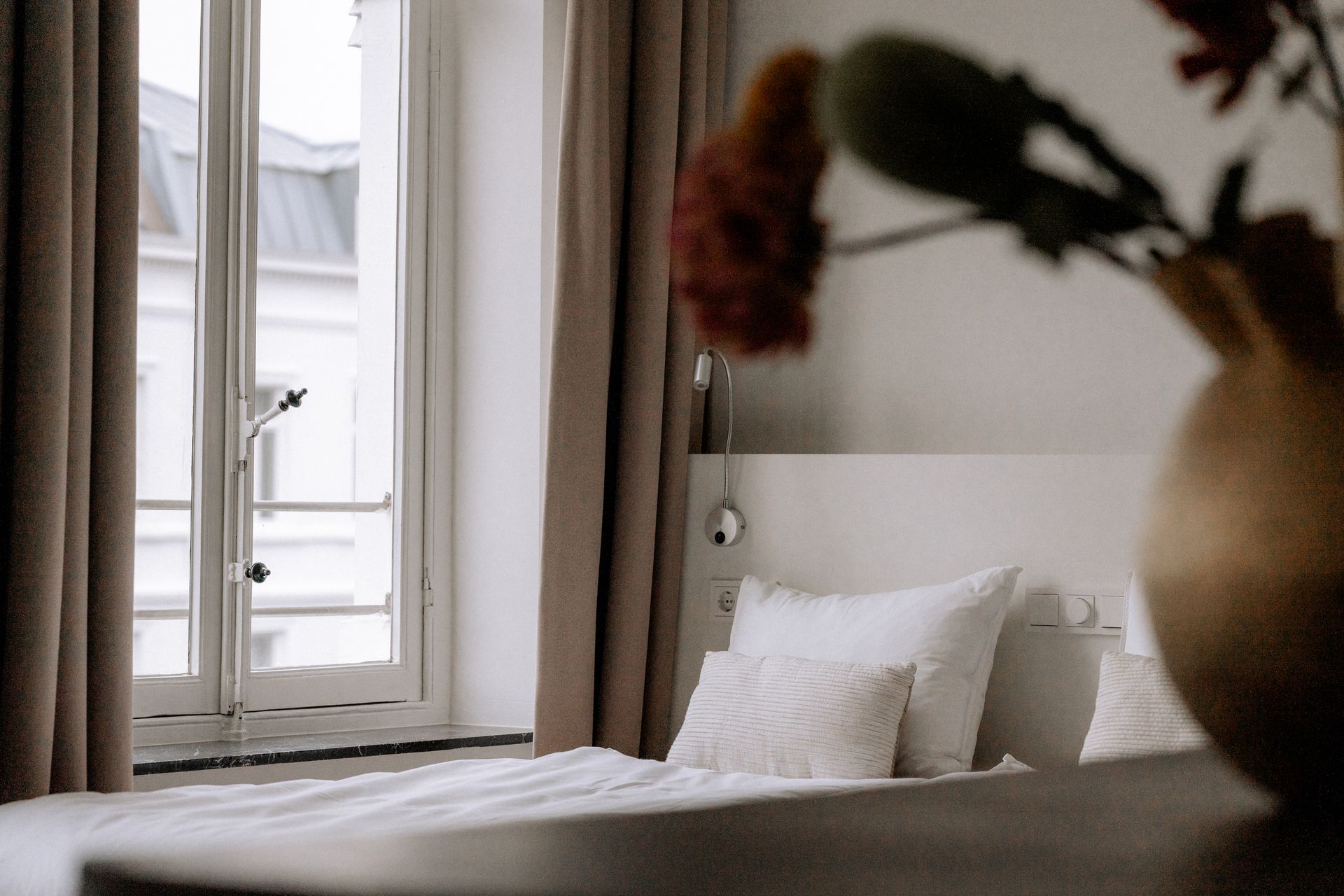 Slaapkamer met wit beddengoed, raam, bruine gordijnen en een vaas met rode bloemen.