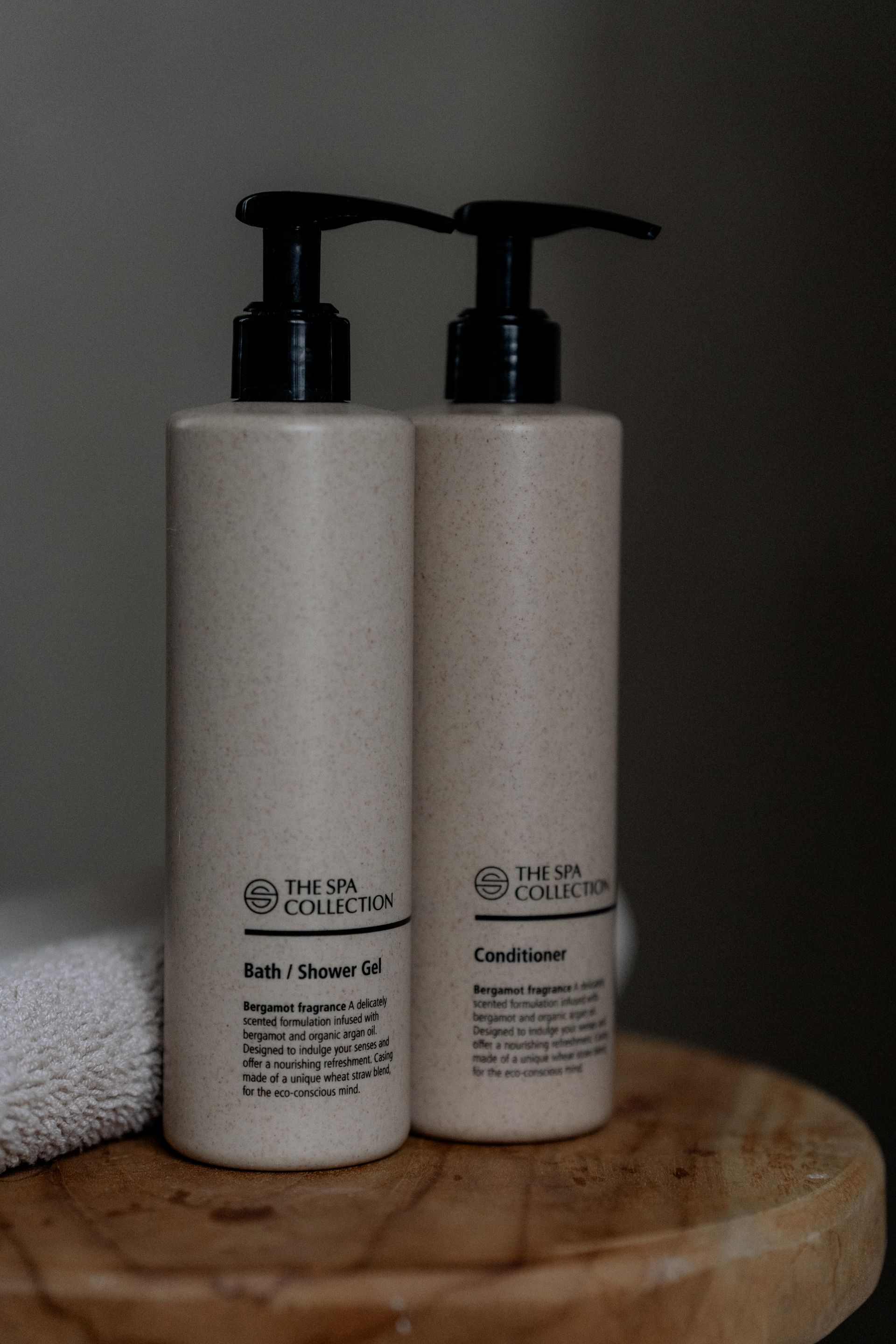 Twee beige flessen shampoo en conditioner met zwarte pompjes staan op een houten krukje, naast een witte handdoek.