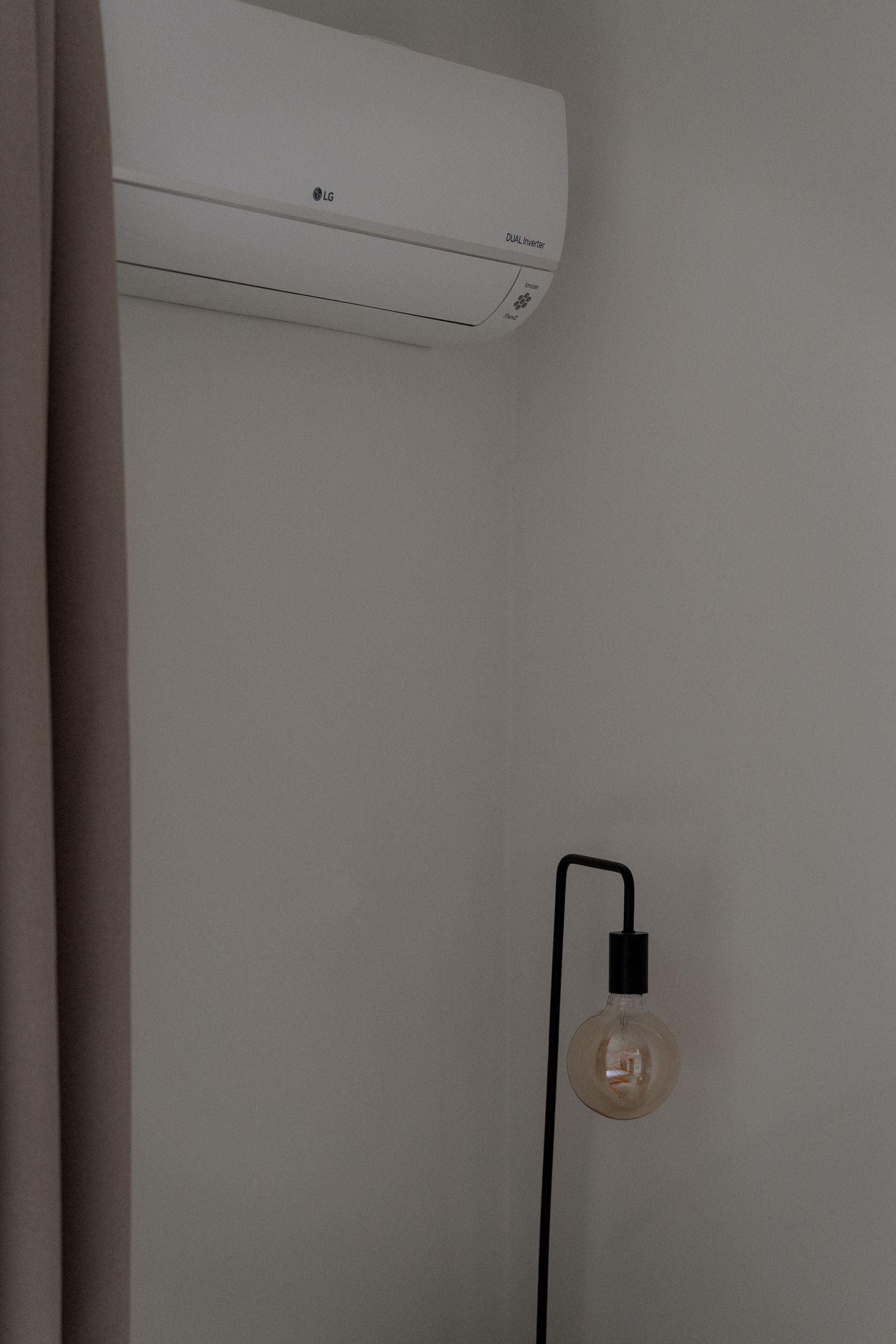 Een witte airconditioningunit boven een zwarte staande lamp met een gloeiende lamp, tegen een lichtgekleurde muur.