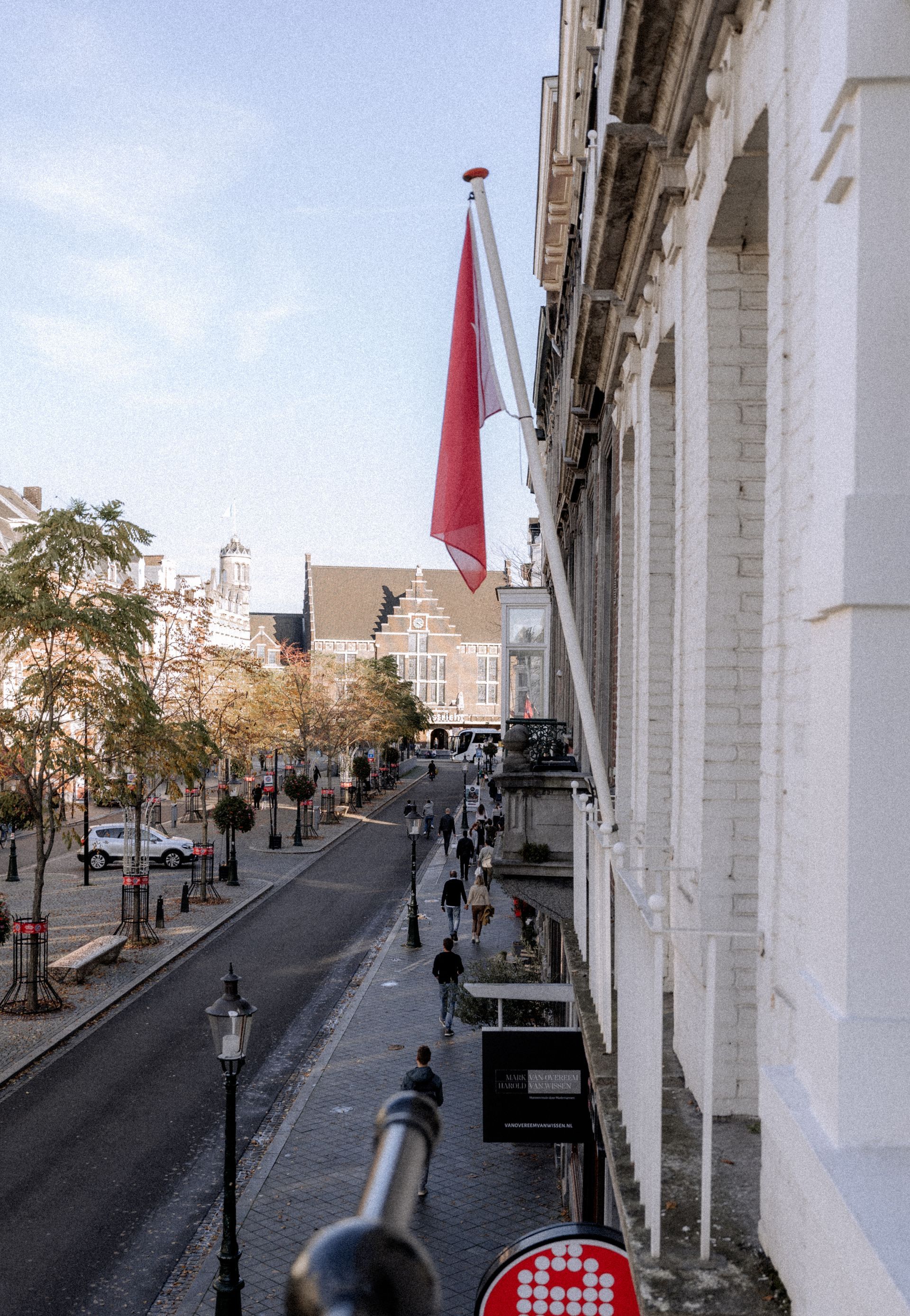 Straatbeeld met een rode vlag, mensen die over de stoep lopen en gebouwen.