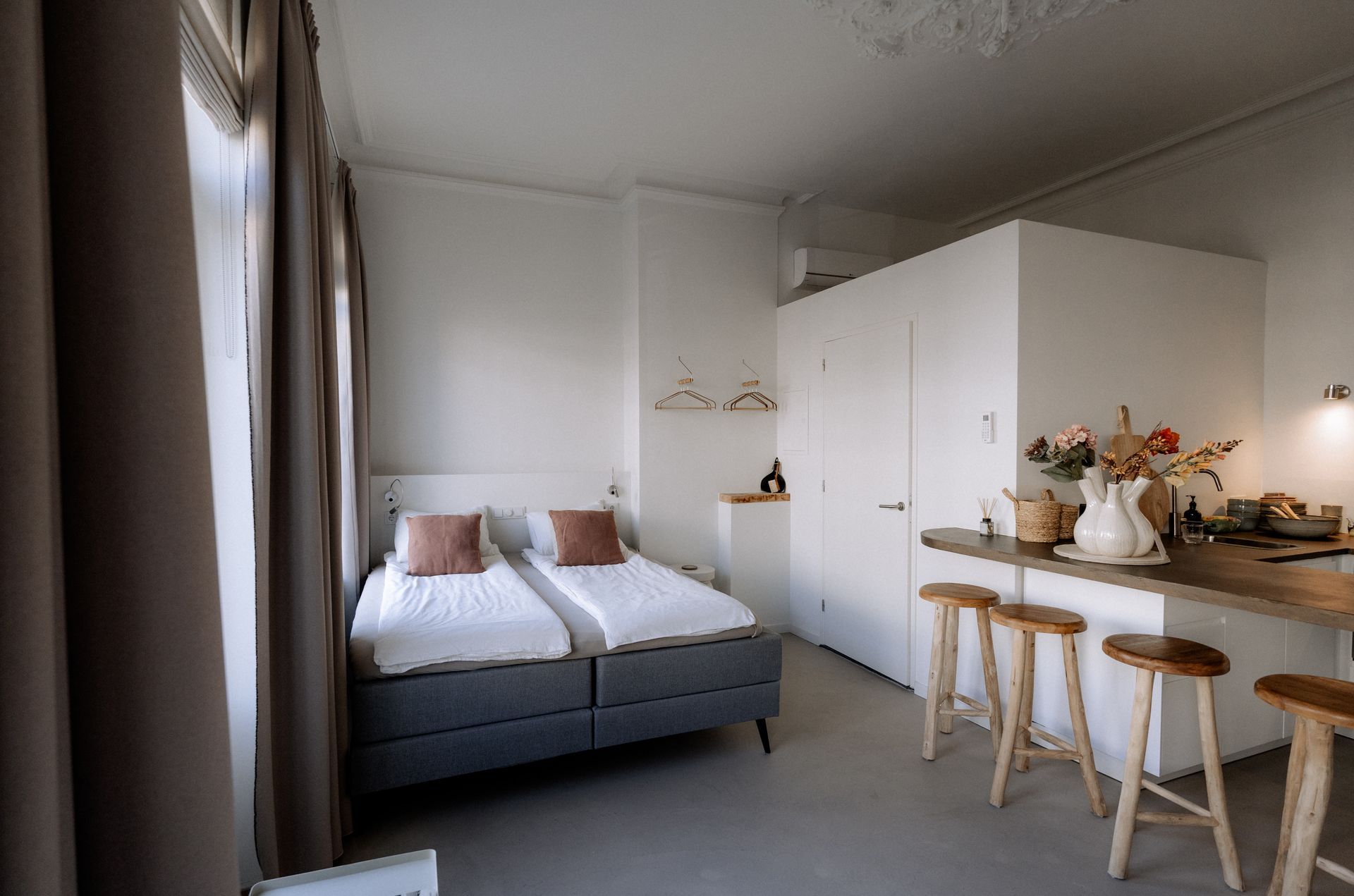 Klein, licht studioappartement met bed, keuken en krukjes. Witte muren, grijs bed, houten aanrechtblad.