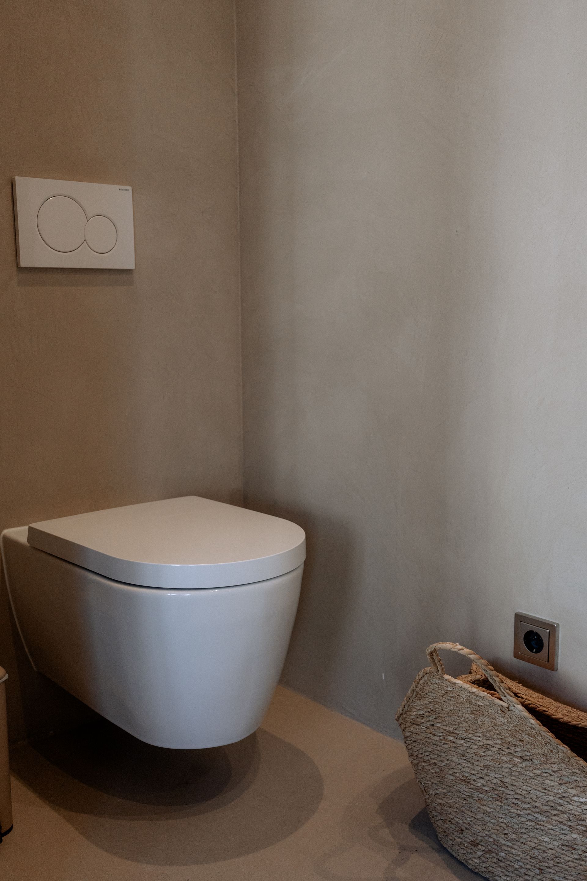 Wit, aan de muur gemonteerd toilet in een lichtgekleurde badkamer. Spoelknop aan de muur erboven. Zeefje in de hoek.