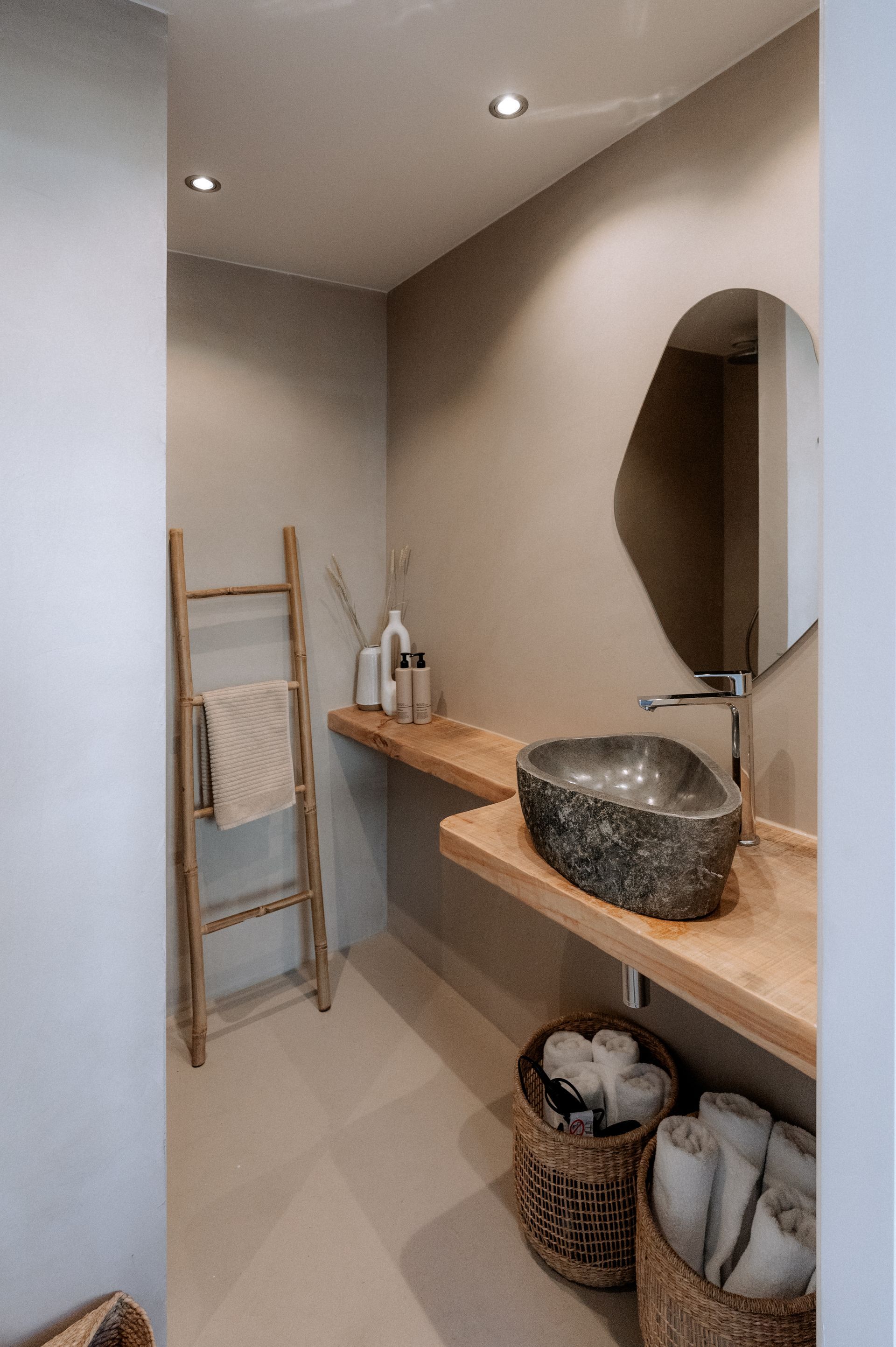 Kleine beige badkamer met houten plank en stenen wastafel. Spiegel, mandjes en handdoekenrek aanwezig.