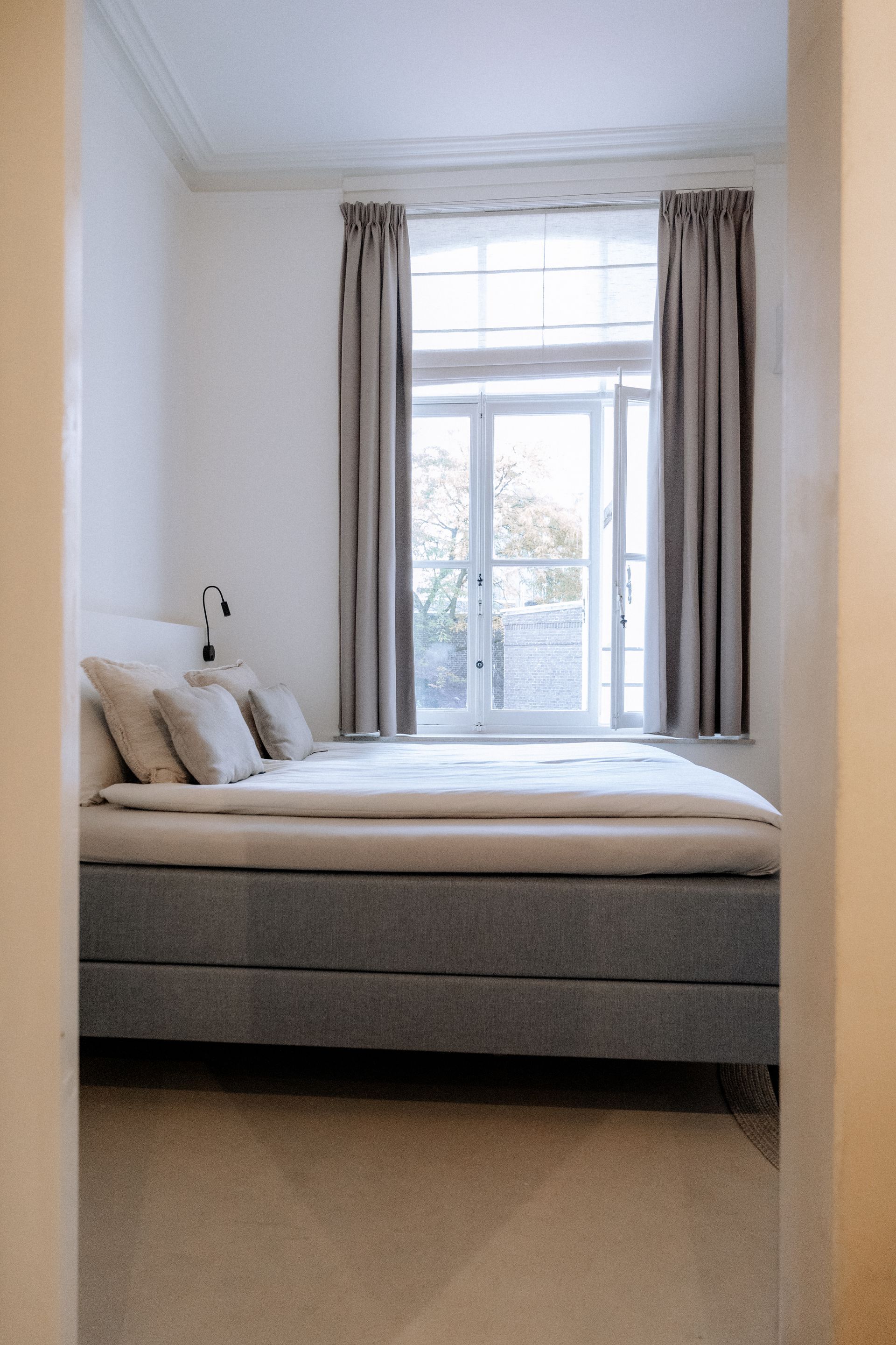 Slaapkamer met bed, kussens en een raam met gordijnen; helder, natuurlijk licht.