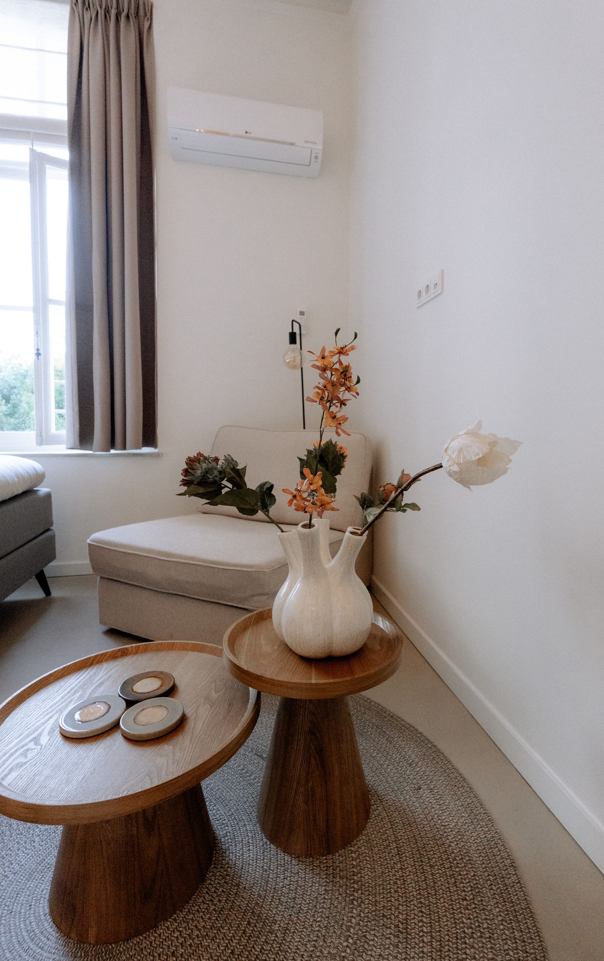 Gezellige woonkamer met fauteuil, salontafels en een vaas met bloemen. Raam met gordijnen en airconditioning.