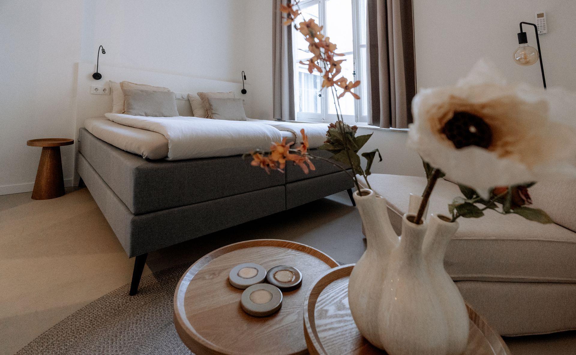 Gezellige slaapkamer met een bed, kleine tafeltjes en een raam. Op de voorgrond staat een vaas met bloemen.