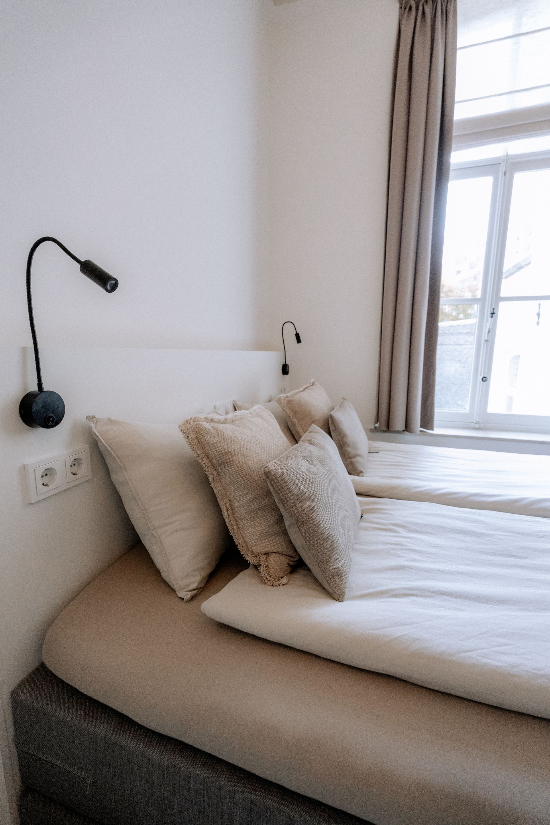 Slaapkamer met twee eenpersoonsbedden, een wit hoofdeinde, beige beddengoed, kussens en een raam met een gordijn.